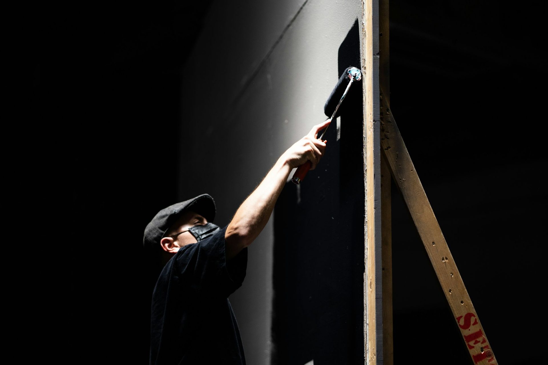 A scenic painter in a cap rolling black paint onto a tall flat in a dark room