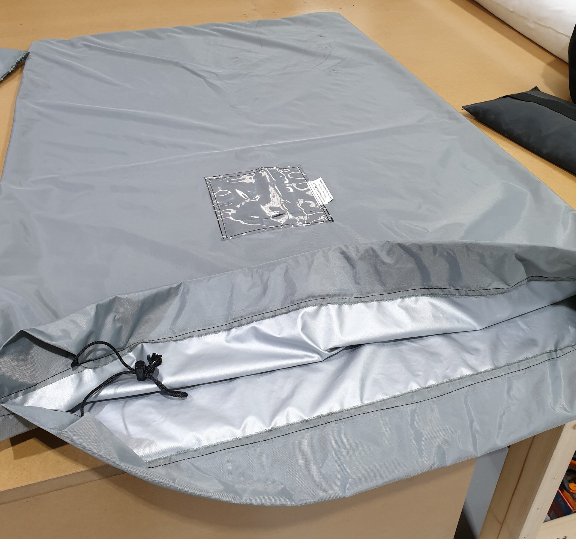 Grey drape bag laid flat on a workshop bench, its drawstring open and a clear label pocket sewn to the face