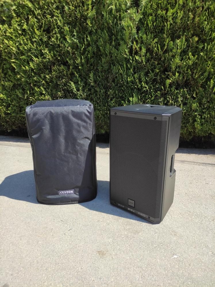 Black fitted speaker cover on one cabinet beside an identical uncovered speaker, standing on a driveway