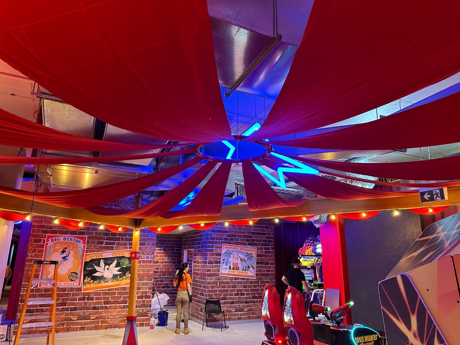 Red fabric swags radiating across a ceiling above arcade machines, with ladders still up mid-install