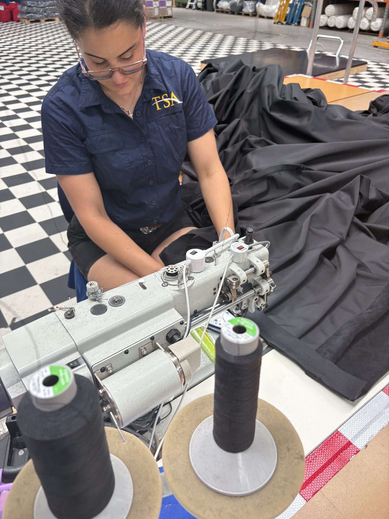A machinist feeding black cloth through an industrial sewing machine, cones of black thread in front