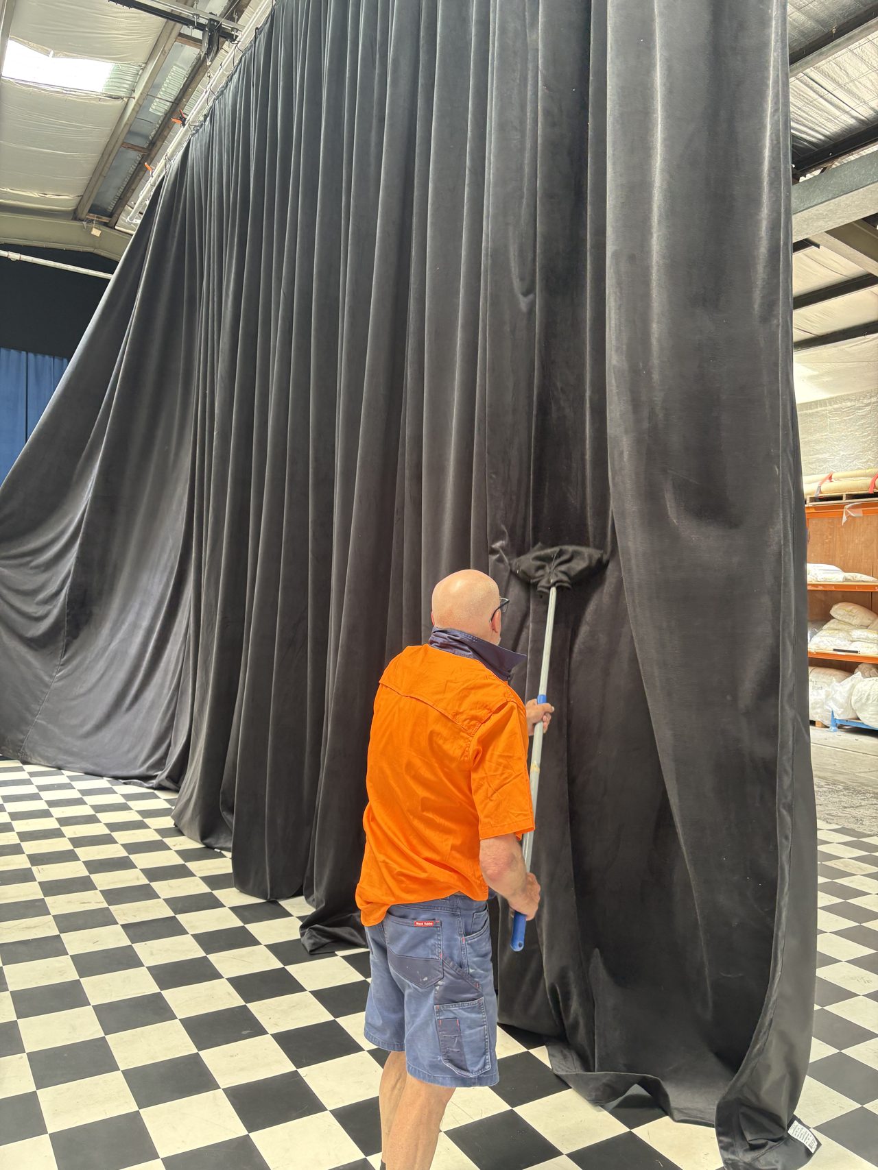 A staff member in hi-vis running a long-handled brush down a tall grey drape in the workshop