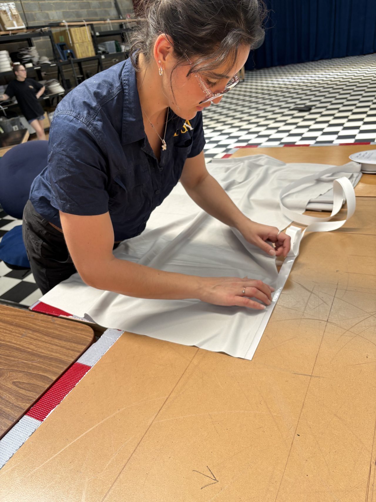 A machinist folding the edge of a white cloth panel on the cutting table, webbing tape beside her