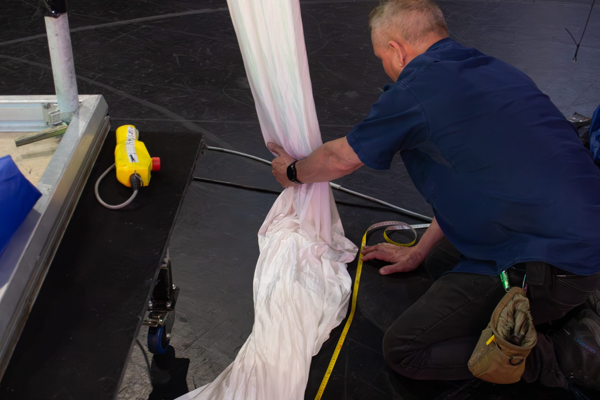 A technician kneeling on a black stage floor, running a tape measure along a length of white cloth