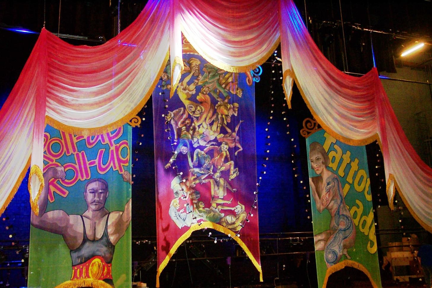 Tall printed banners with circus-poster artwork hung between red and white swags under arena lighting