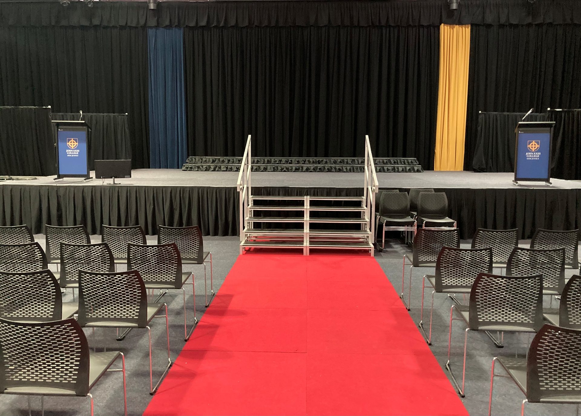 Pleated black skirting along the front of a school hall stage, a stair unit and red carpet leading up to it, black drapes behind