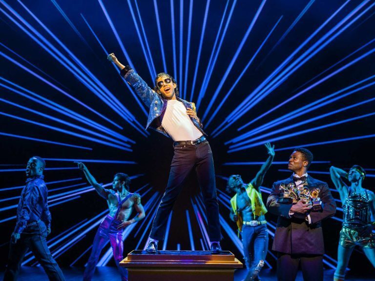 Performer as MJ in a sequinned jacket, arm raised centre stage against radiating blue beams of light in MJ the Musical