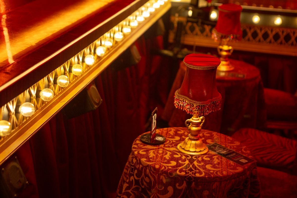 Red velvet booth seating and a fringed table lamp beneath the gilded balcony of the Moulin Rouge! set