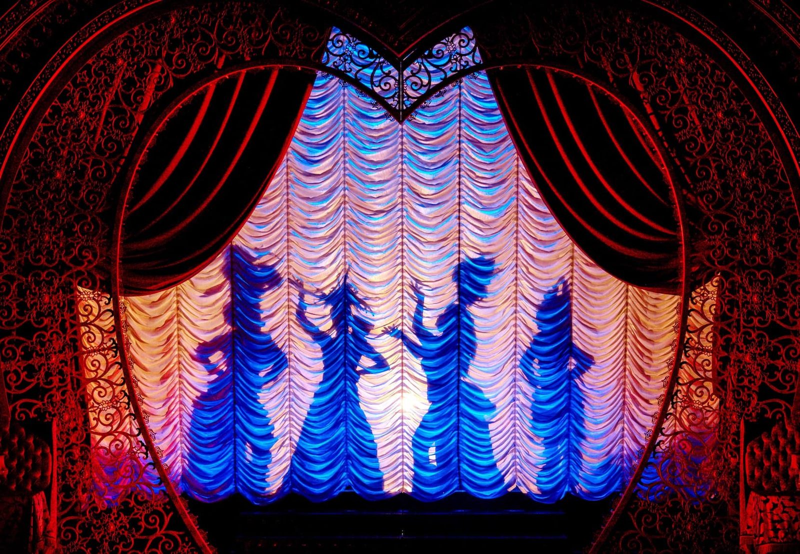 Swagged red drapery framing a lit blue festoon curtain, with dancers in silhouette on the Moulin Rouge! stage