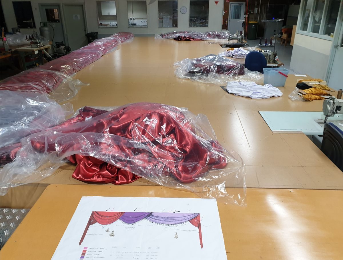 Red velvet drapery in progress on long workshop tables, with a coloured drawing of the drape laid beside it