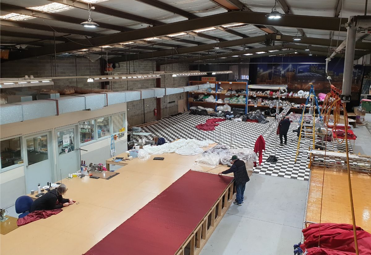 A long run of red fabric laid along a cutting table in a wide workshop, makers working at the far end
