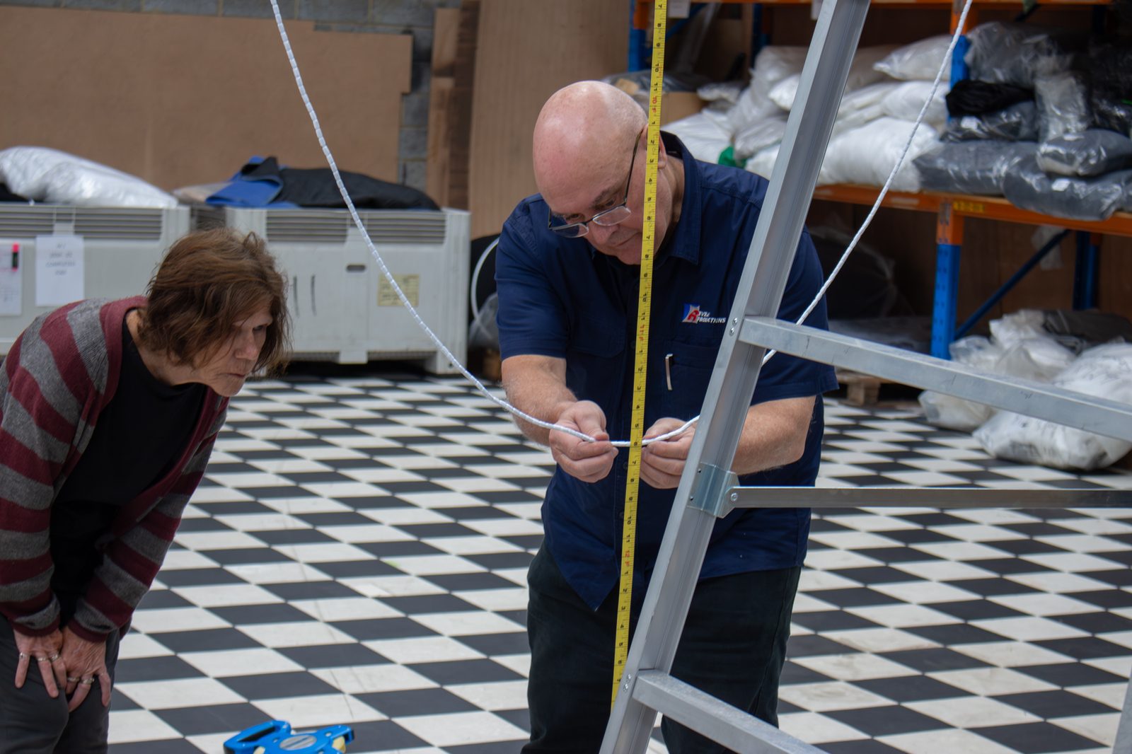A maker reading a tape measure on the workshop floor while a colleague leans in to check it