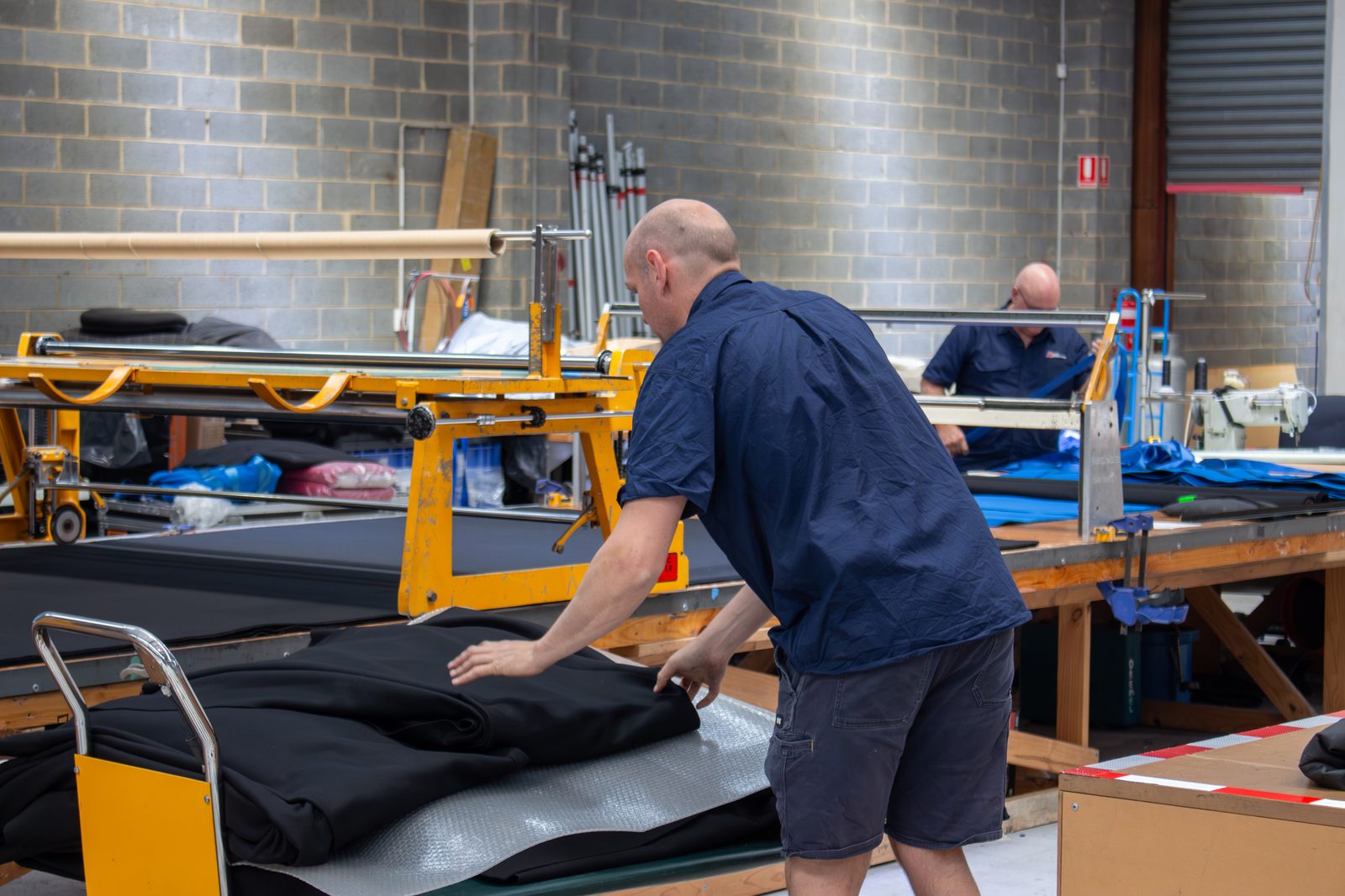 A machinist smoothing black drape fabric at the workshop cutting table, with the fabric spreader behind