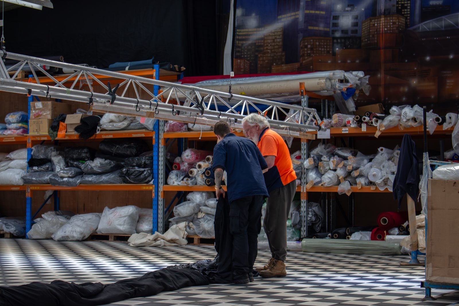 Two workshop staff handling a long black drape in front of the fabric store shelves