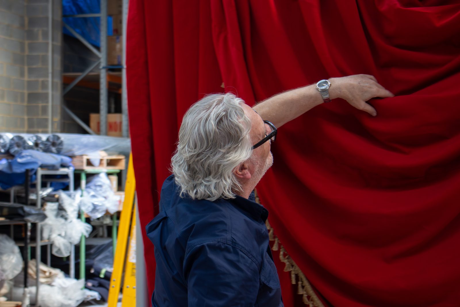 A technician reaching up to check a red velvet drape with gold fringe in the workshop