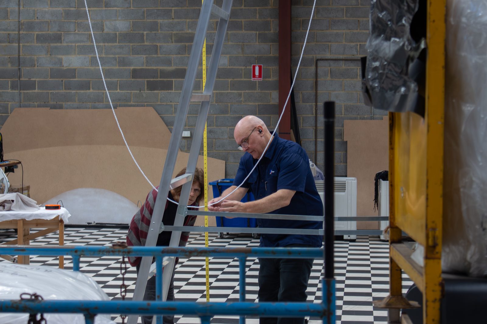 A maker running a tape measure down a ladder on the workshop floor while a colleague checks the reading