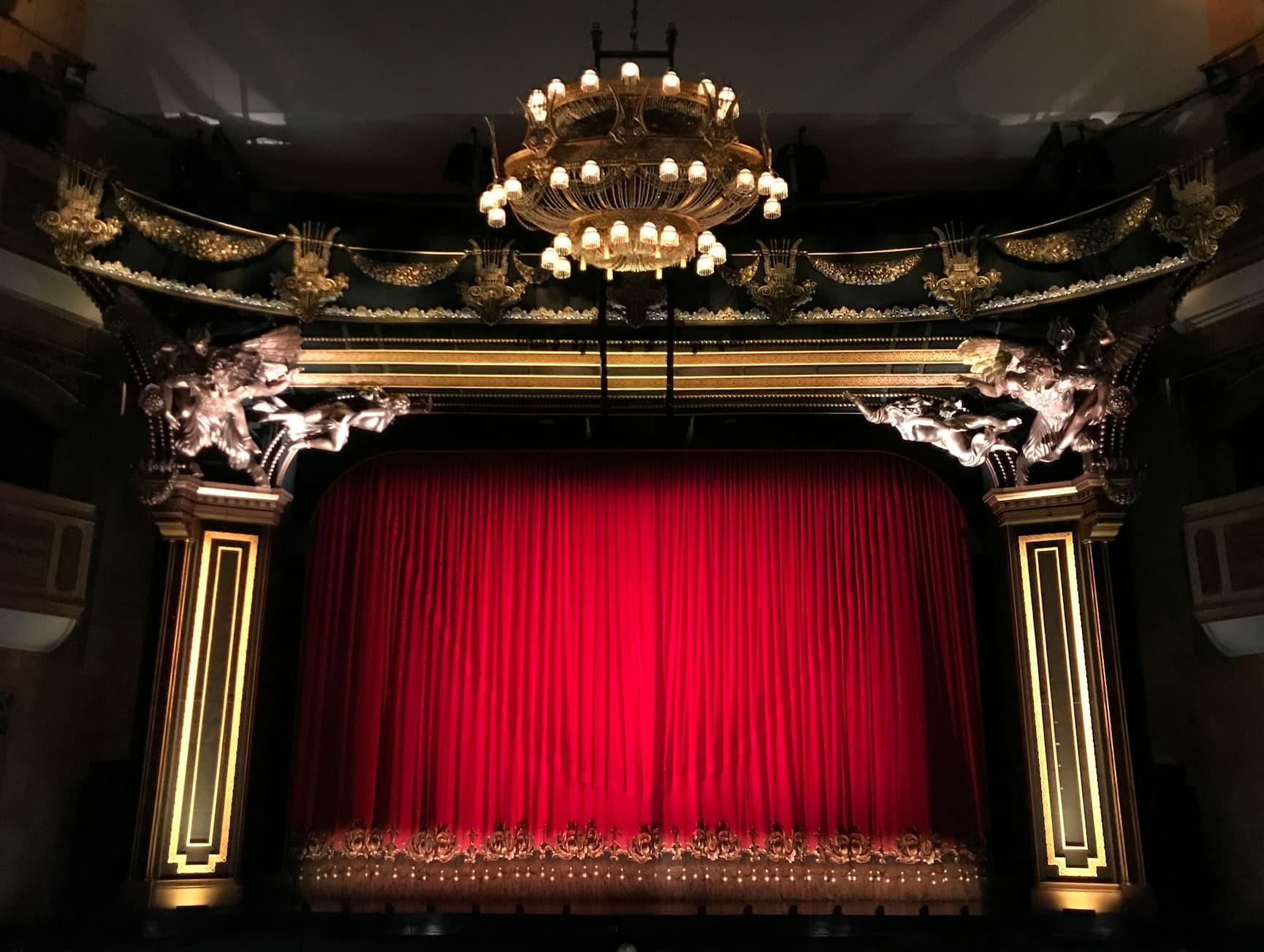 A red velvet house curtain hanging in an ornate theatre proscenium beneath a chandelier