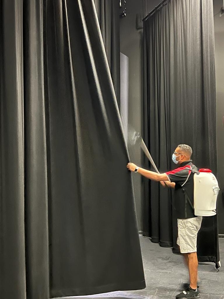 A technician with a backpack sprayer misting a hung black stage curtain in a venue