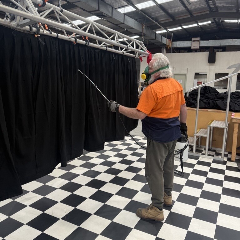 A technician in hi-vis and a respirator spraying black drapes hung from a truss in the workshop